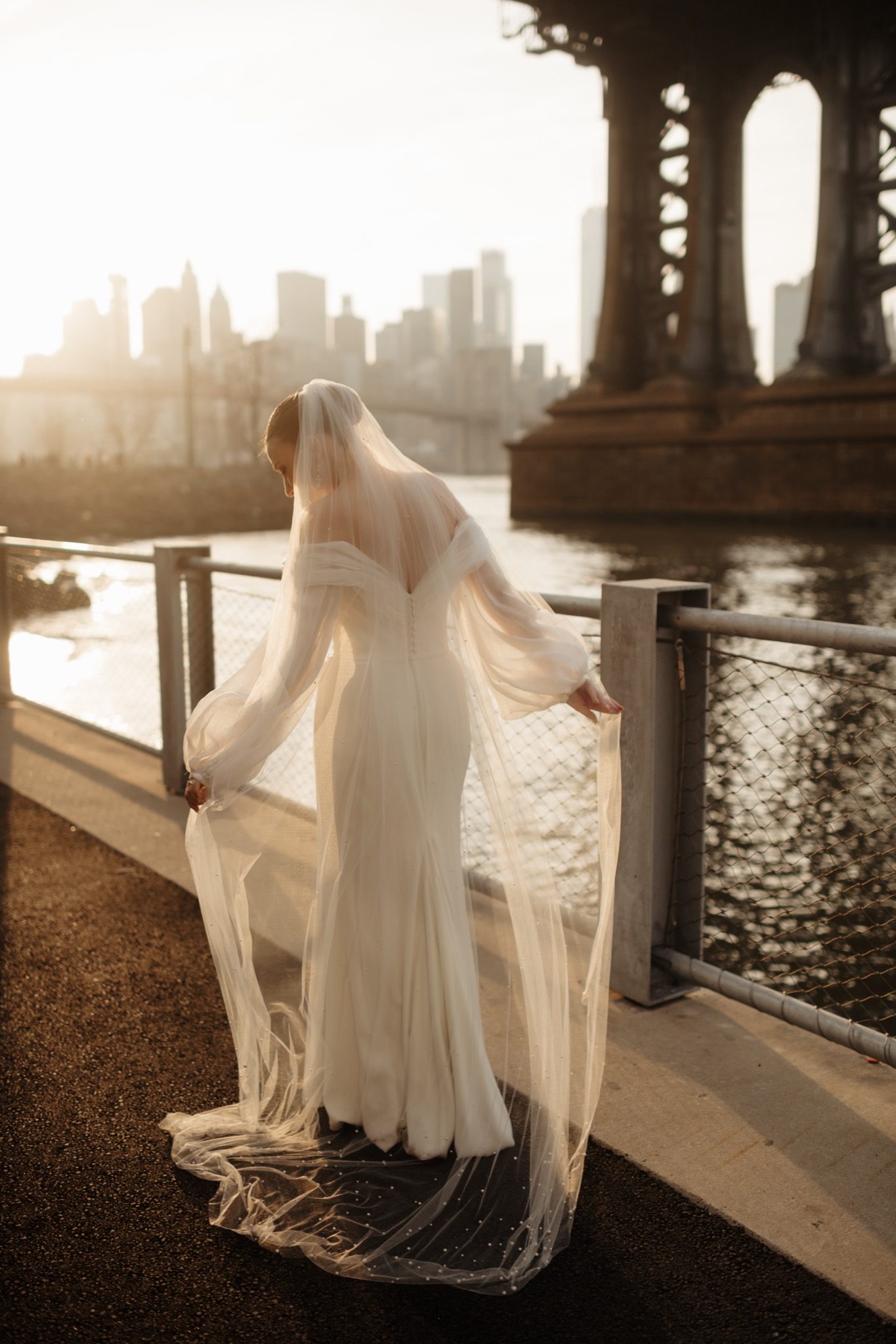 Bride with her long veil at the East River railing with the Manhattan skyline at golden hour, Brooklyn NYC | New York wedding photographer Tonie Christine